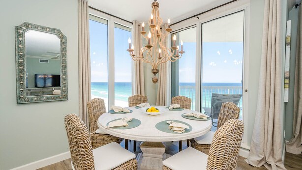 A dining area with a round table, wicker chairs, a chandelier, and a view of the ocean. - FIRST WEEK OF AUGUST JUST OPENED! (Destin)
