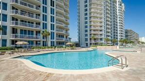 A swimming pool area with lounge chairs and umbrellas, surrounded by multi-story buildings with balconies. - FIRST WEEK OF AUGUST JUST OPENED! (Destin)