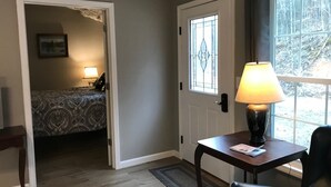 Une pièce avec un lit, une table avec une lampe, une porte avec une fenêtre et une vue sur les arbres à l’extérieur.