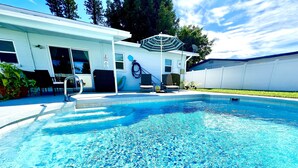 A swimming pool with clear blue water, a covered patio area with outdoor furniture, and a white building with a satellite dish. - Private Heated  Pool House 6 miles from the Beach (Bradenton)