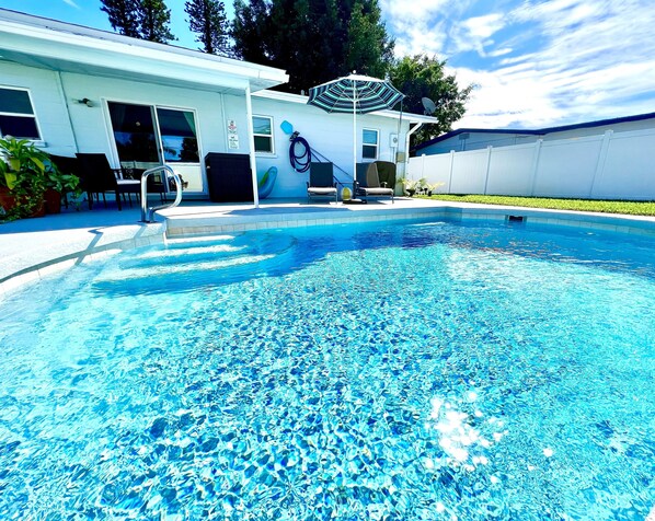 Une piscine à l’eau d’un bleu cristallin, un patio couvert avec des meubles d’extérieur, et un bâtiment blanc surmonté d’une antenne parabolique.
