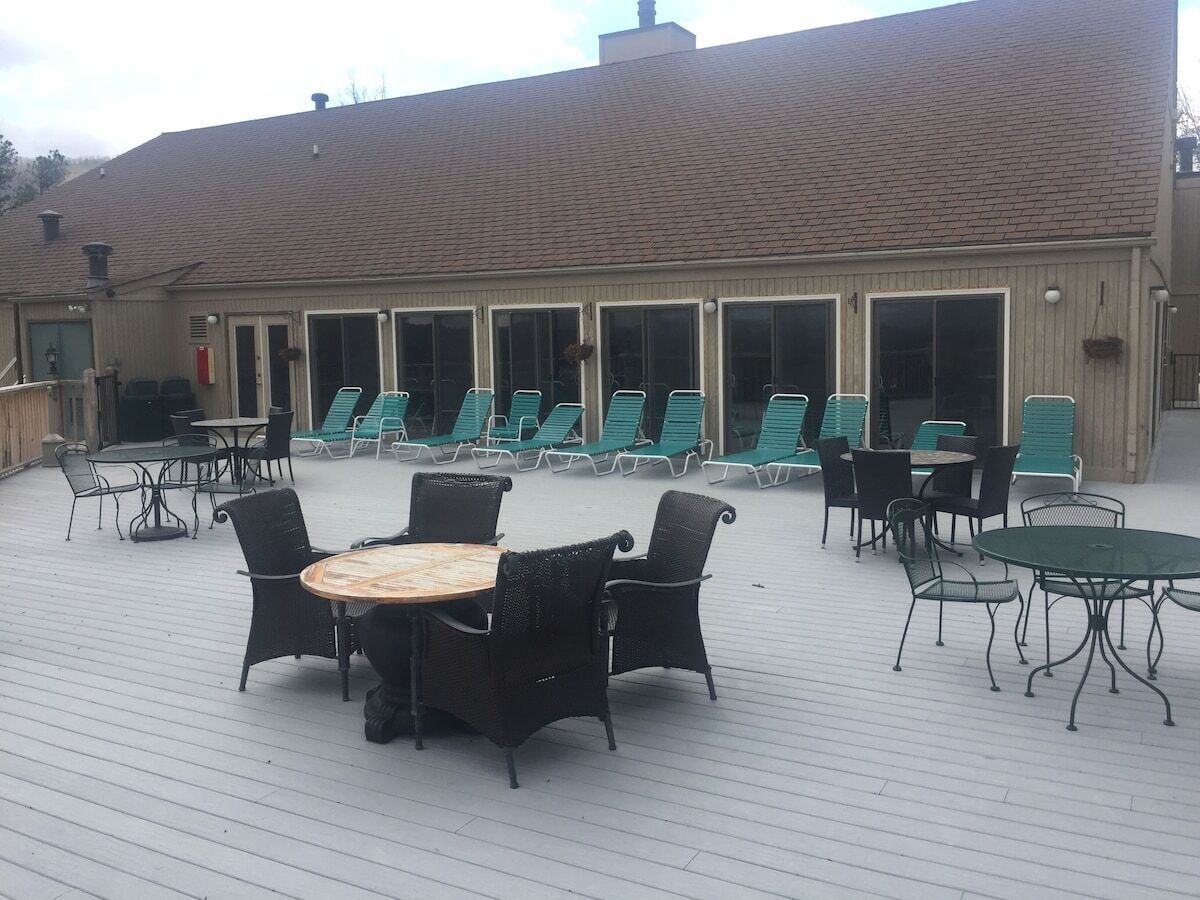 Une terrasse extérieure couverte, dotée d’une table ronde et de chaises, entourée de fauteuils et bordée d’un bâtiment aux grandes fenêtres.