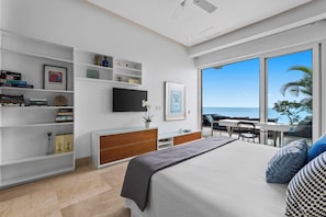 A modern bedroom with a bed, a TV, a desk, and a balcony with a view of the ocean.