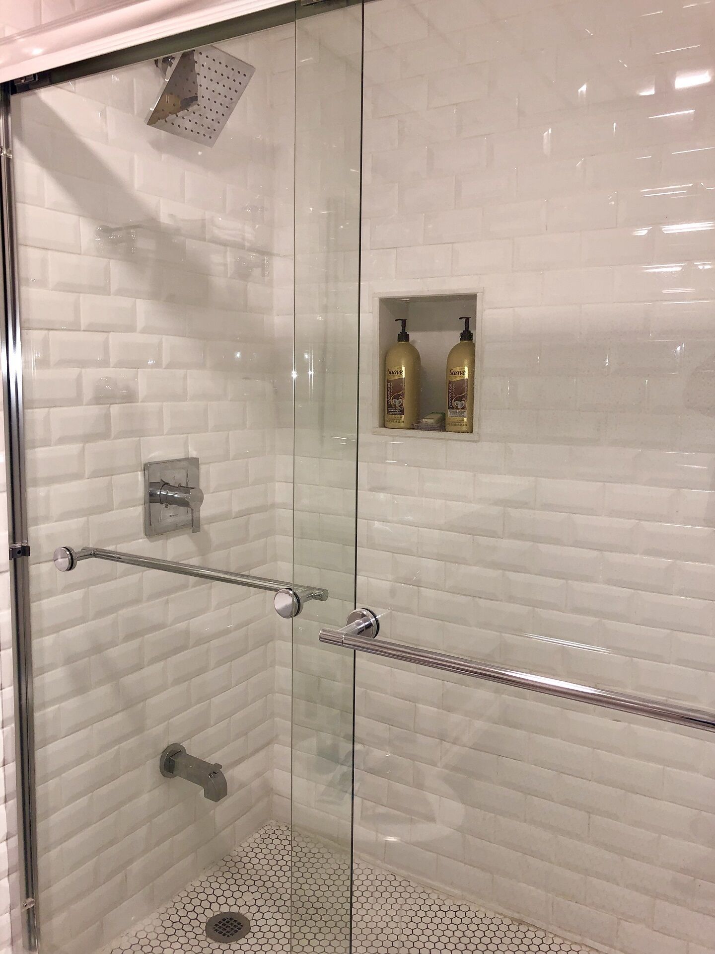 A glass-enclosed shower with white tiled walls, a glass door, and a metal showerhead. There are two bottles of shampoo on a shelf above the shower.