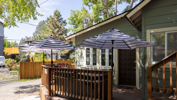 A wooden deck with two striped umbrellas, a wooden fence, and a house with a dark front door. - Contemporary Chic │West Side │Walk Everywhere │Hot Tub │One Block to River (Bend)