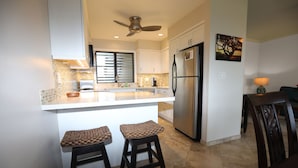 A modern kitchen with a central island, two bar stools, a refrigerator, and a ceiling fan. - Paradise found-Ocean Front 2 Bdr-Bath-Lanais—Primary bedroom faces ocean FULL AC (Kapaa)