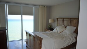 A bedroom with a wooden bed, a nightstand, a lamp, and a view of the ocean through large sliding doors.