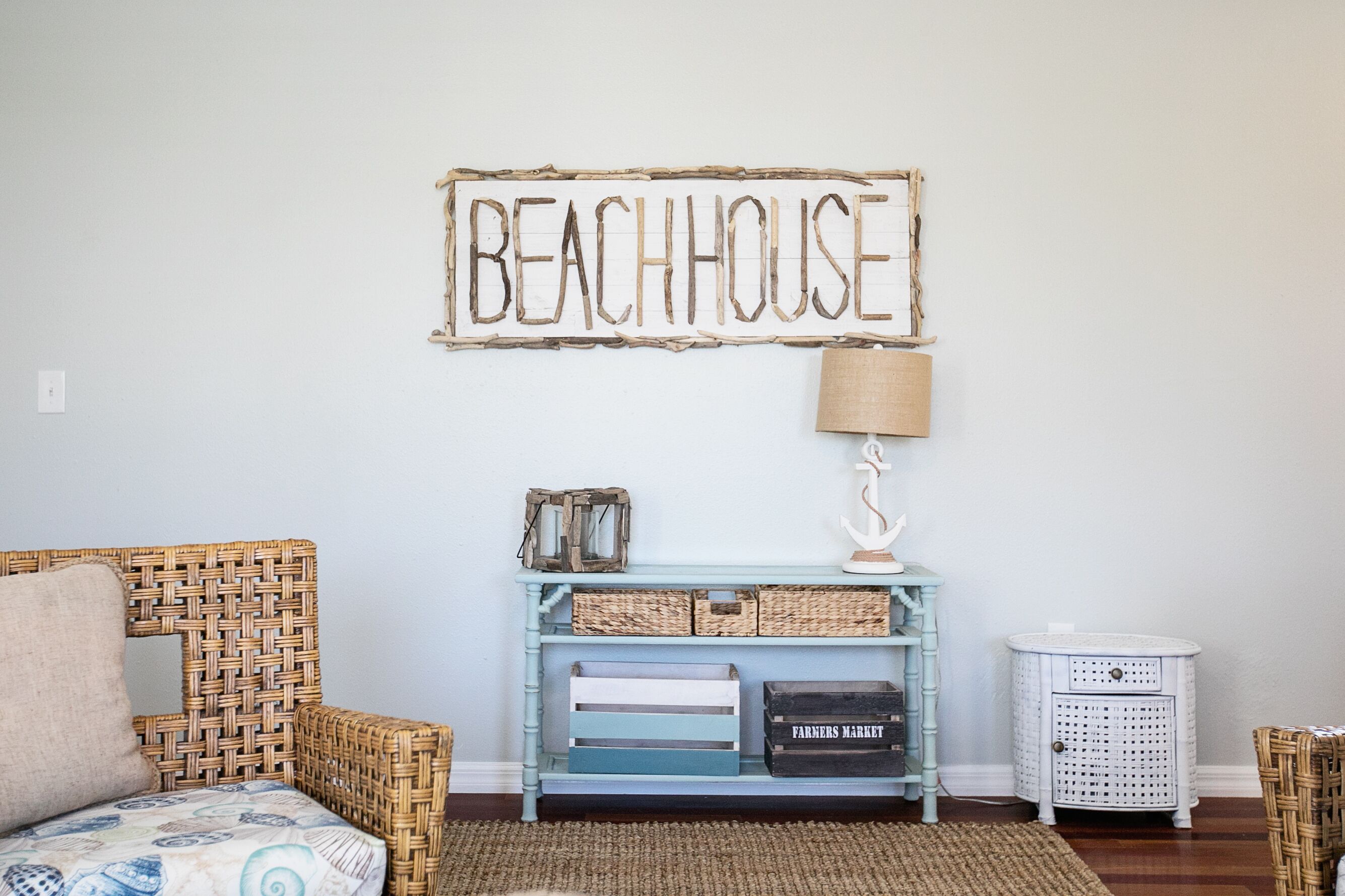 Une pièce avec un fauteuil en osier, une console bleue agrémentée de paniers et un panneau mural sur lequel on peut lire « BEACHHOUSE ».