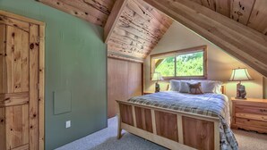 Une chambre avec un plafond en bois, un lit recouvert d’une couverture à carreaux, une table de chevet avec une lampe et une fenêtre donnant sur un paysage verdoyant.