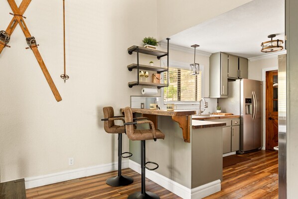 A kitchen with wooden flooring, a built-in island with bar stools, stainless steel appliances, and a pendant light. - Lux Cabin Retreat: Outdoor Soaking Tubs, Sauna, Firepit, Game Room for kids (Lake Arrowhead)