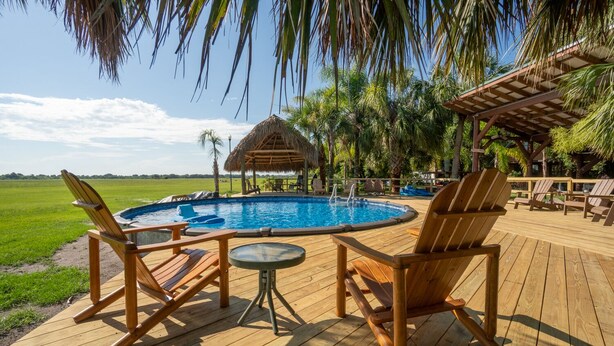 Terraza de madera con piscina, sillas de descanso y un pabellón con techo de paja.