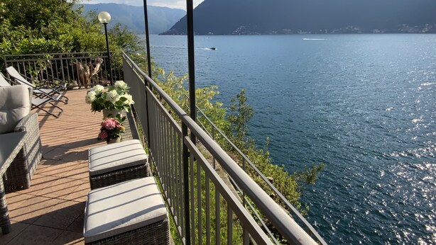 A balcony with wicker furniture and potted plants, overlooking a lake with mountains in the background. - Outdoor Veranda w Lakefront Views, Lake Access & Swimming Dock (COMO)