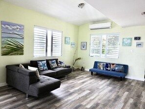 Sala de estar com sofá modular cinza, um sofá azul e piso de madeira. Há duas janelas com persianas brancas, um ventilador de teto e obras de arte nas paredes.