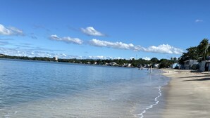 A beach with clear water, a sandy shore, and a distant shoreline with buildings and trees.