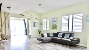 A spacious living room with a grey sectional sofa, a blue armchair, and a wooden floor. There are large windows with blinds, a ceiling fan, and a door leading to another room.
