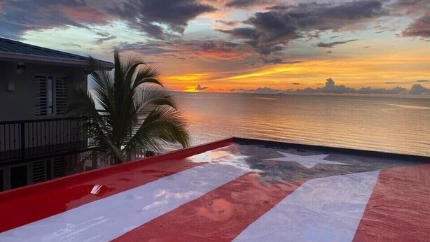 A balcony with a view of the sunset over the water, palm trees, and a building with balconies.