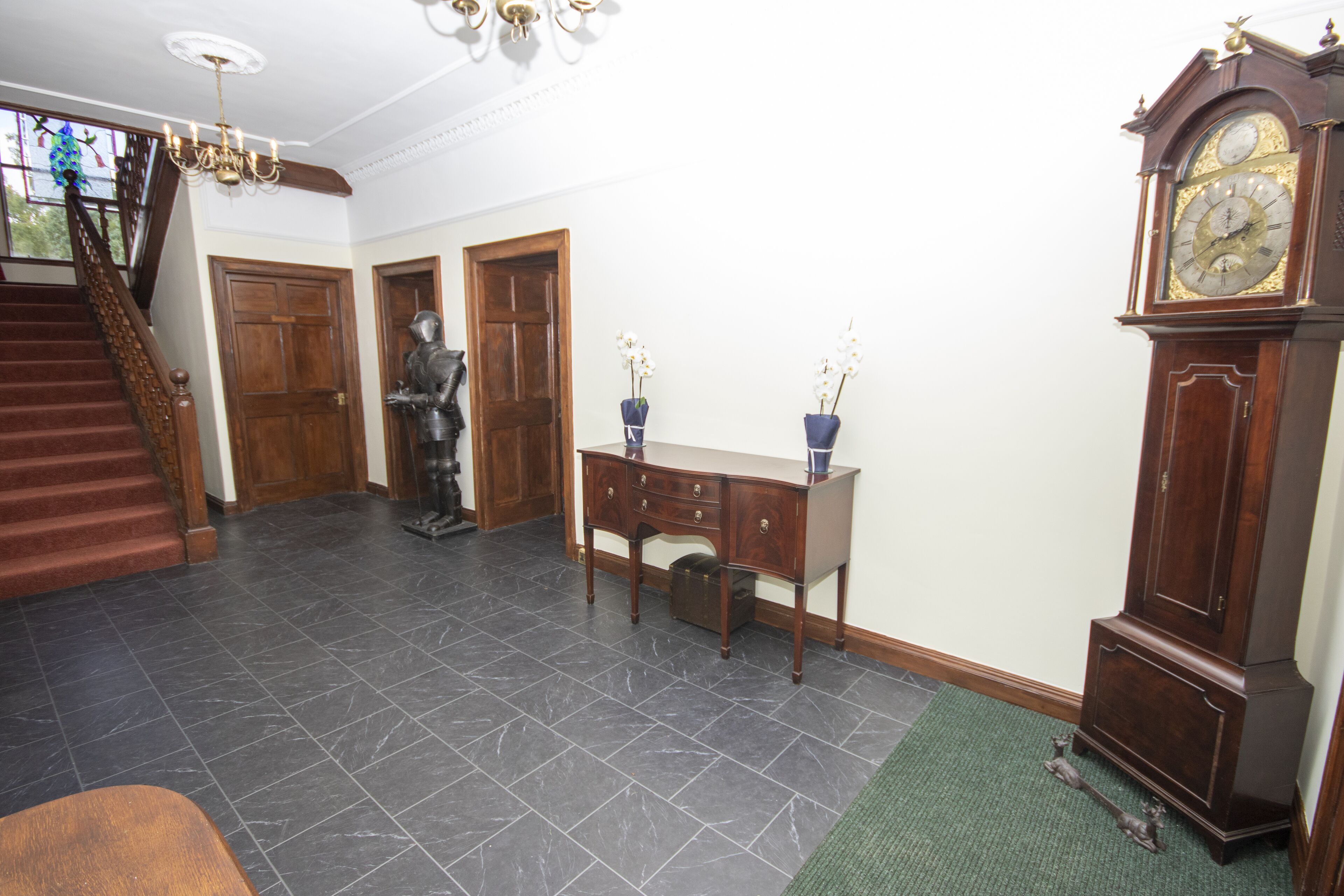A spacious hallway with a grand staircase, a wooden desk with two blue vases, and a large wall-mounted clock.