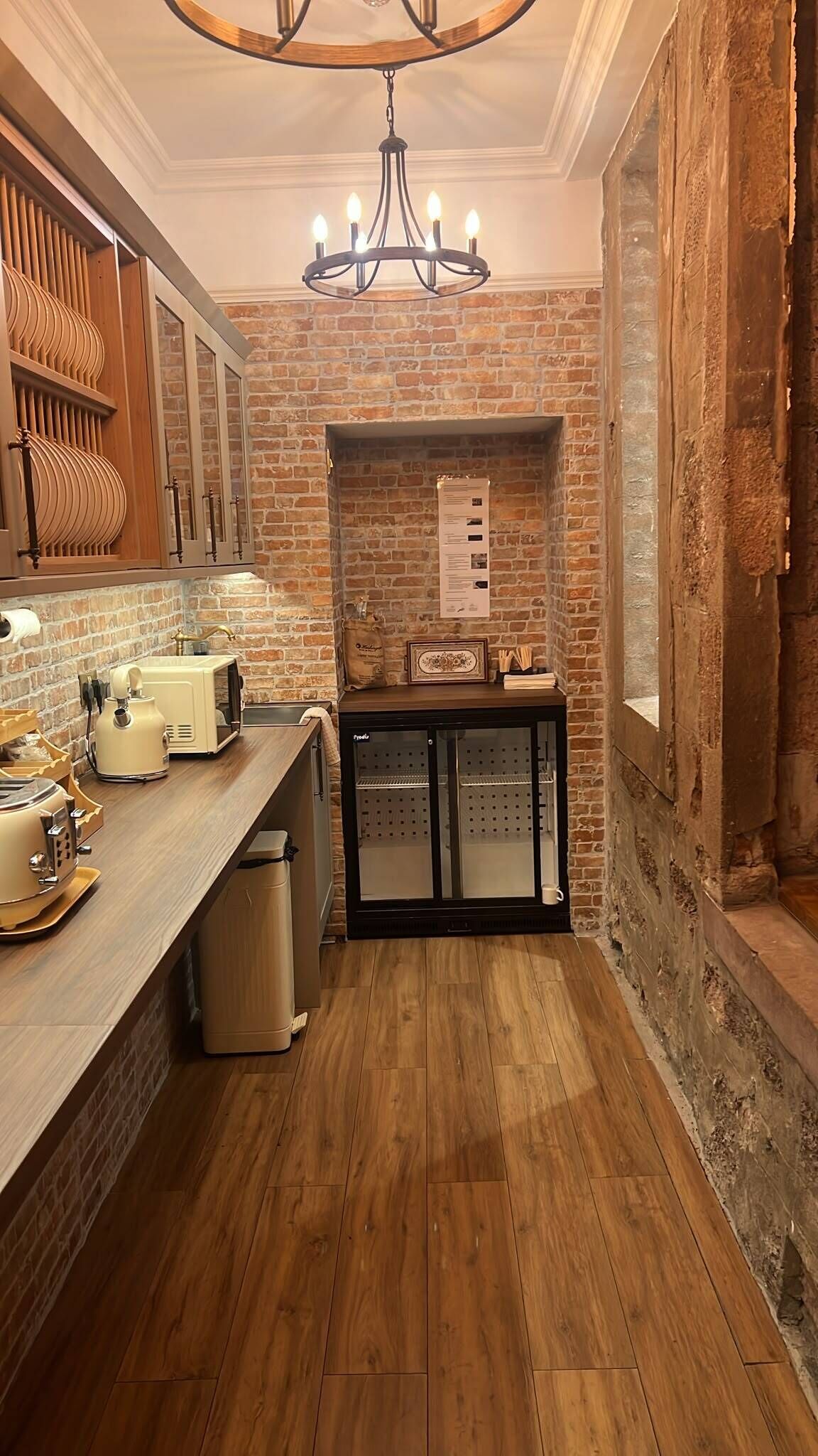 A kitchen with wooden flooring, a brick wall, a chandelier, and a refrigerator.