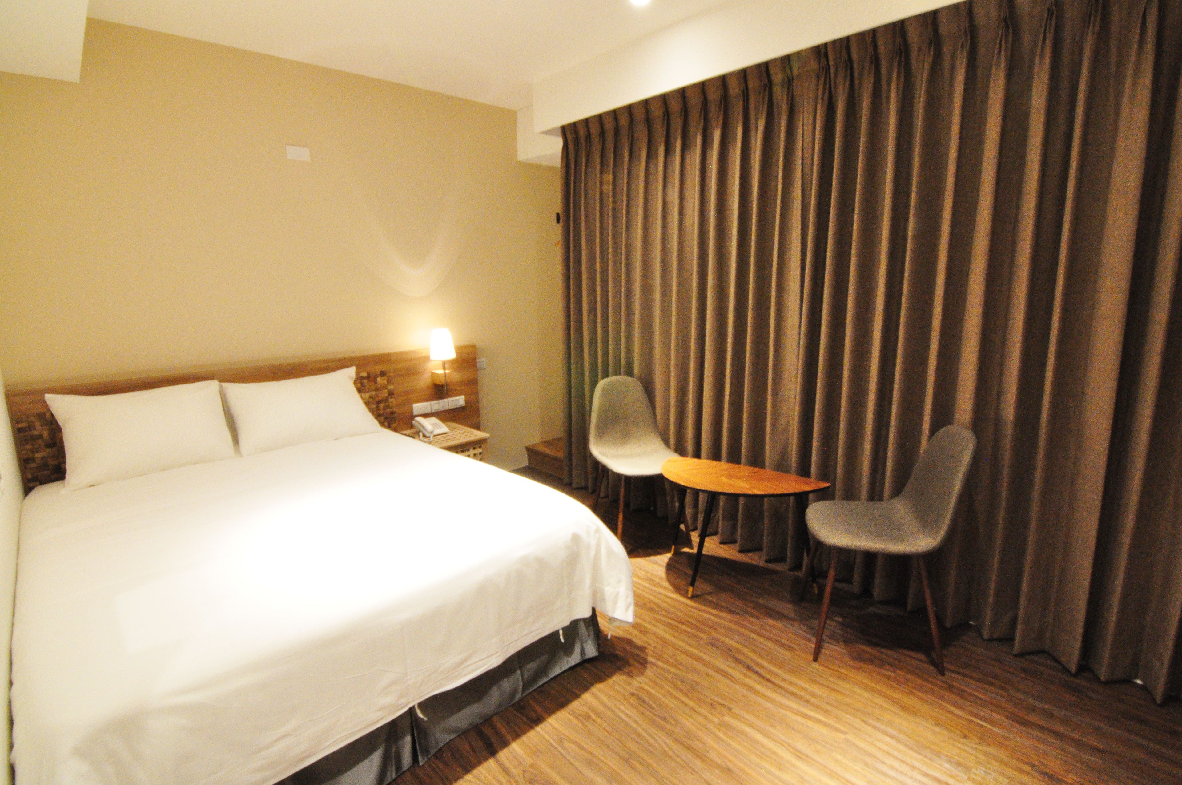 A hotel room with a large bed, a desk with a lamp, a chair, and a curtain.