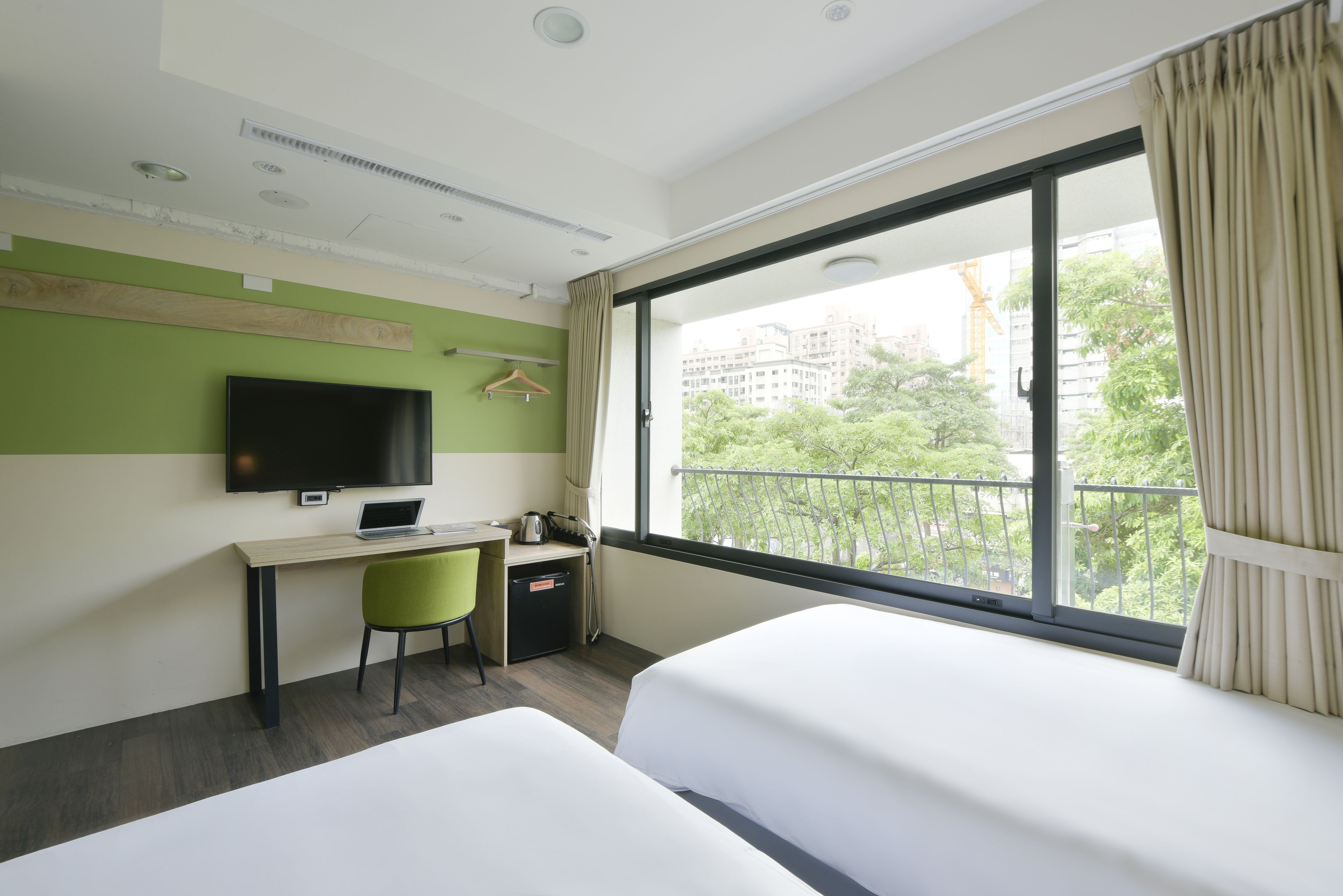 A hotel room with two beds, a desk with a laptop, a television, and a view of the city through large windows.