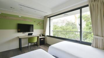 A hotel room with two beds, a desk with a laptop, a television, and a view of the city through large windows.