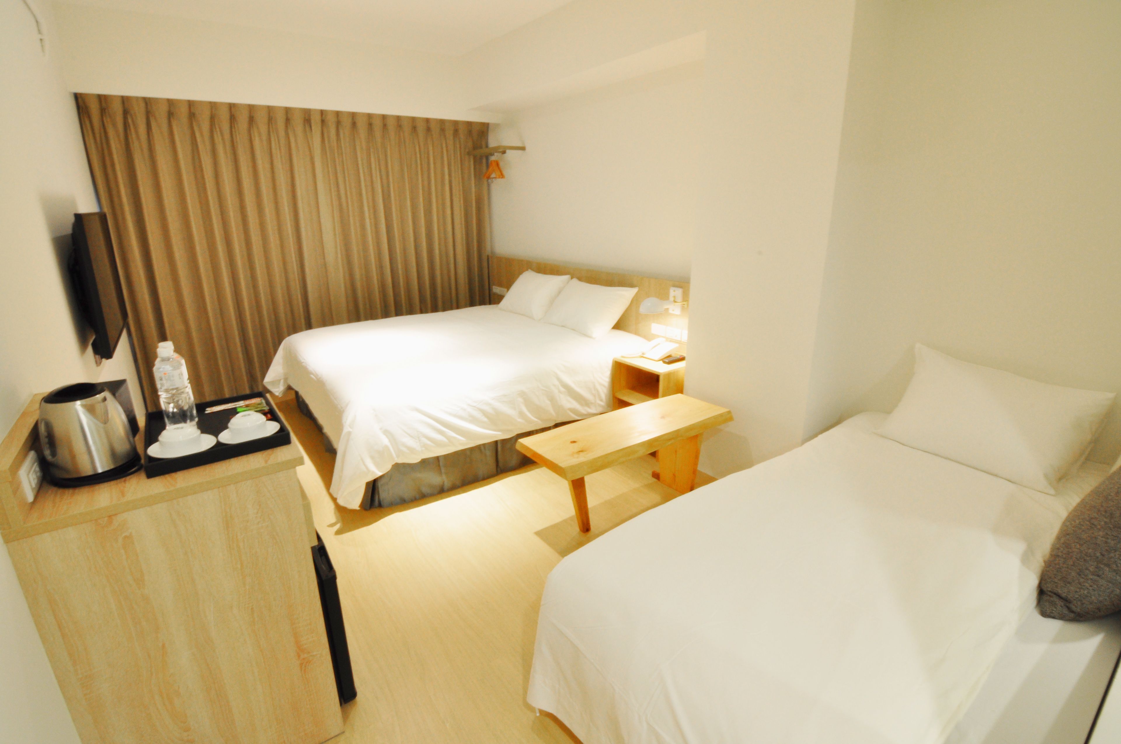 Habitación de hotel con dos camas, un escritorio de madera con hervidor y botella de agua, una mesita de noche con lámpara y un ventanal con cortinas.