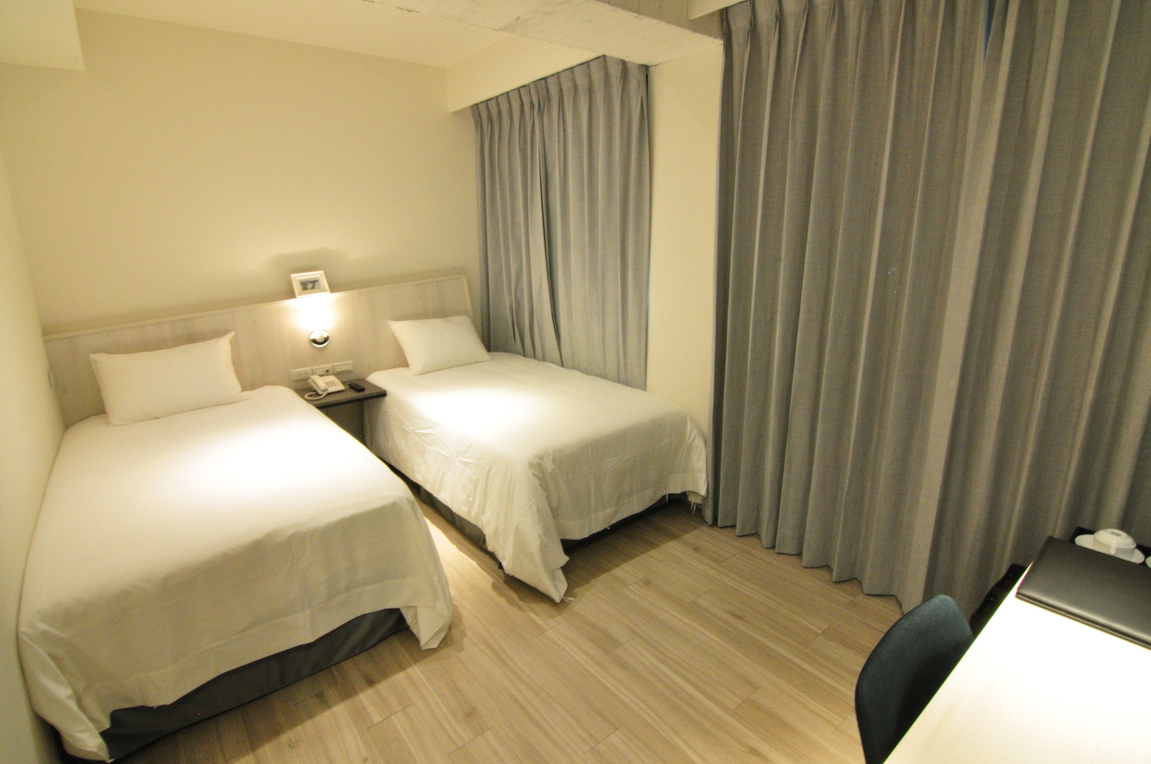 A hotel room with two single beds, a desk with a chair, a nightstand with a lamp, and a large window with curtains.
