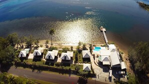Luchtbeeld van een kustresort met meerdere accommodaties in bungalowstijl, een steiger die in zee uitloopt, en een helder uitzicht op de oceaan.