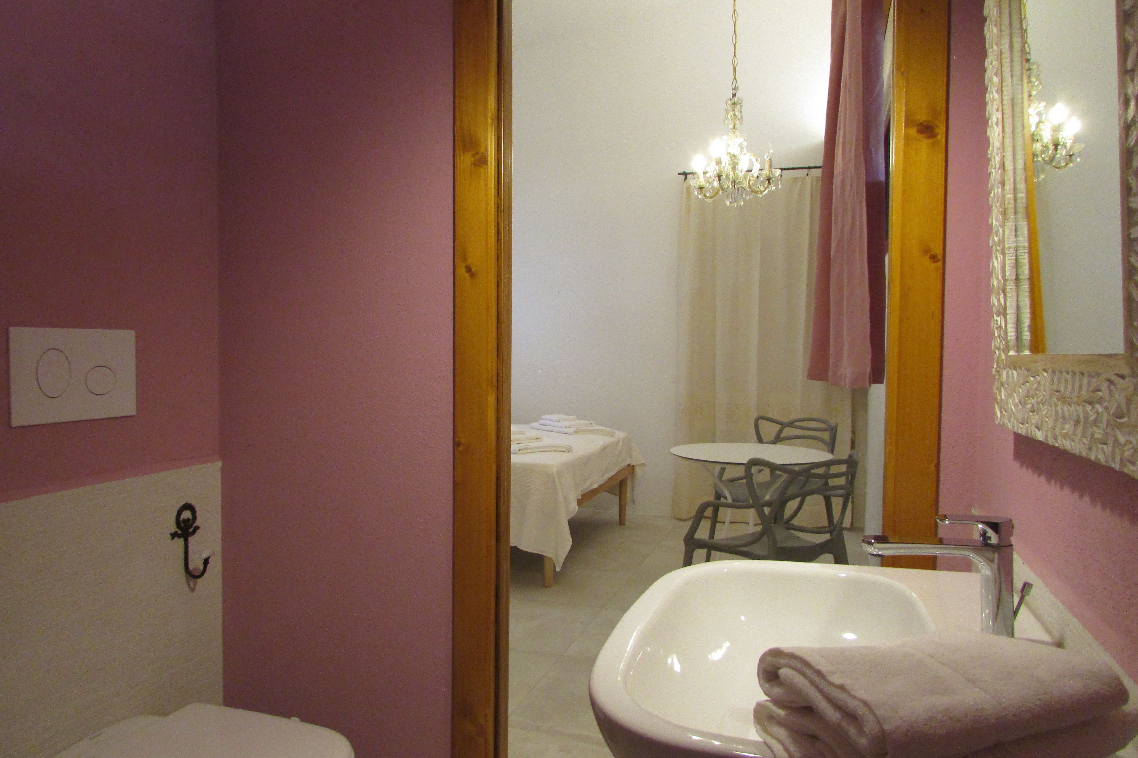 Un bagno con lavandino, WC e uno specchio. Nella stanza adiacente si trova un tavolo e delle sedie.
