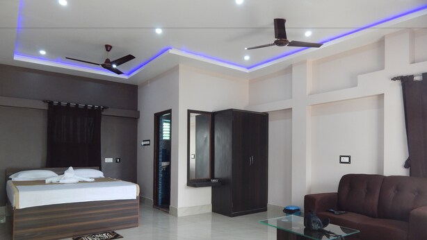 A modern hotel room with a bed, a sofa, a glass coffee table, a wardrobe, and a ceiling fan. - Samriddhi Banquet Garden & Resort (Baharampur)