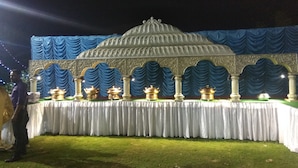 An outdoor event setup with a stage featuring a decorative backdrop and a table with lit lamps. - Samriddhi Banquet Garden & Resort (Baharampur)