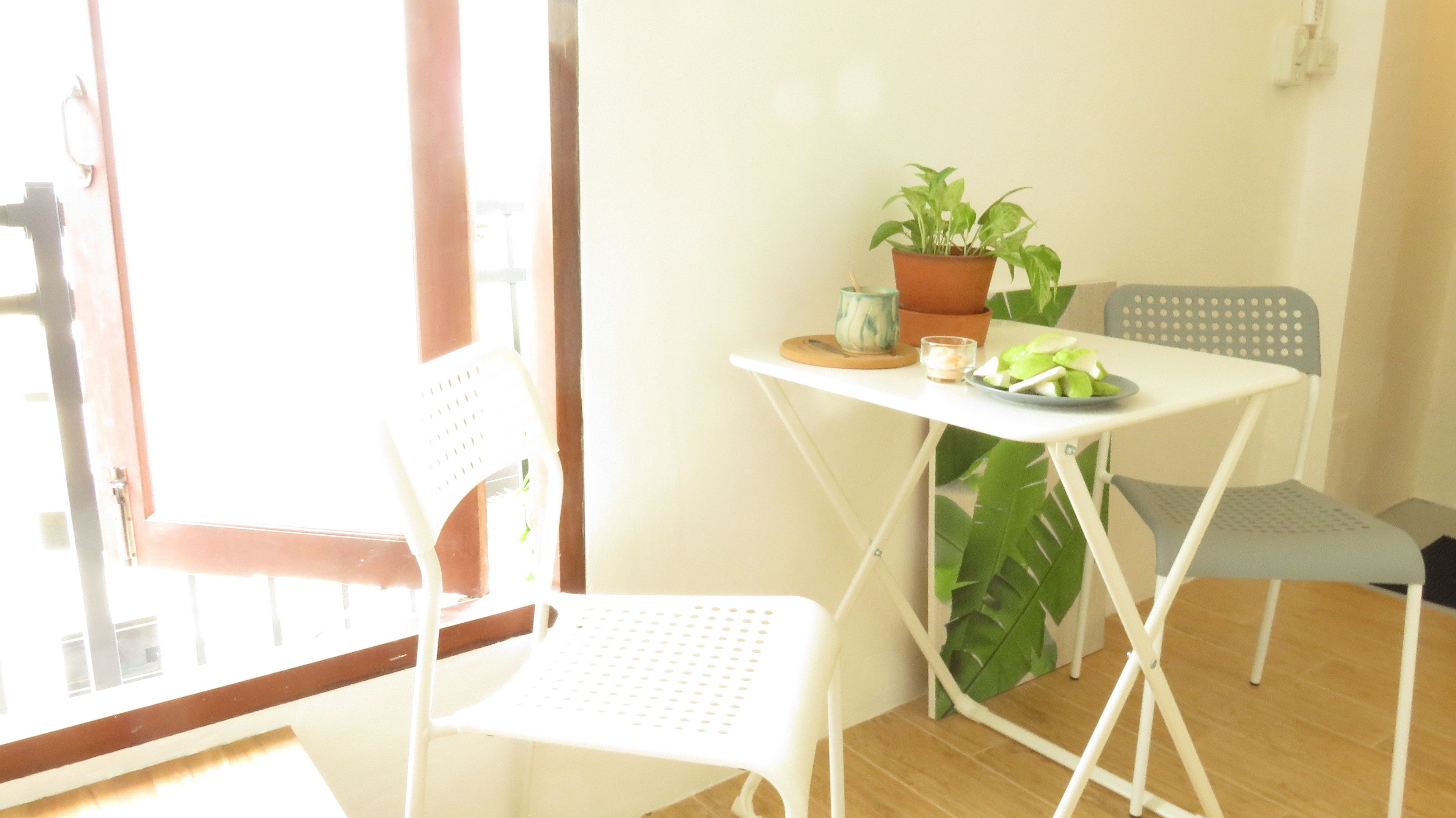 A small, bright room with a white table and two white chairs. There is a potted plant on the table and a window with a view of the outside.