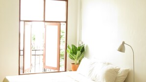 Een kamer met een bed, een bloempot, een bureaulamp en een raam met uitzicht op groen buiten.