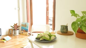 Een kamer met een houten vloer, een witte stoel, een tafel met een bord groene appels, een potplant, een ingelijst schilderij en een kleine tafel met een kop en schoteltje.