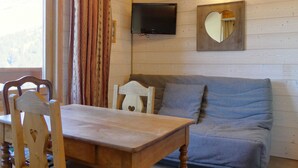 A cozy room with a wooden table, two chairs, a grey sofa, a television mounted on the wall, and a heart-shaped mirror. - Quiet studio with balcony, parking and pets allowed (Méribel)