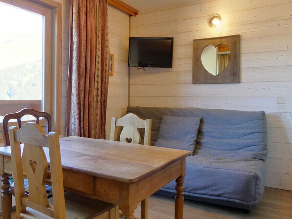 A cozy room with a wooden table, two chairs, a grey sofa, a television mounted on the wall, and a heart-shaped mirror. - Quiet studio with balcony, parking and pets allowed (Méribel)