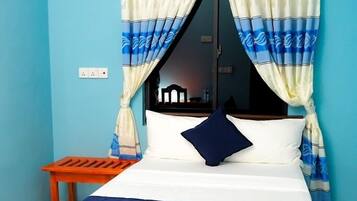 Een bed met houten frame, beddengoed in wit en blauw, en een blauw kussen. Er staat een klein houten tafeltje naast het bed.