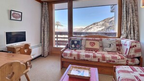 Um quarto acolhedor com uma escrivaninha de madeira, televisão de tela plana, sofá com almofadas estampadas e vista para uma paisagem montanhosa coberta de neve através de grandes janelas.