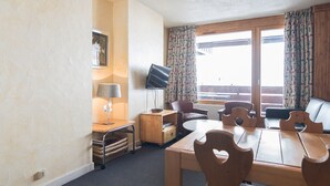 A room with a wooden dining table, chairs, a TV, a lamp, and a window with curtains.