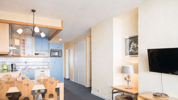 A compact living space with a kitchenette, a dining area with wooden chairs, a flat-screen TV, and a small table with a lamp.