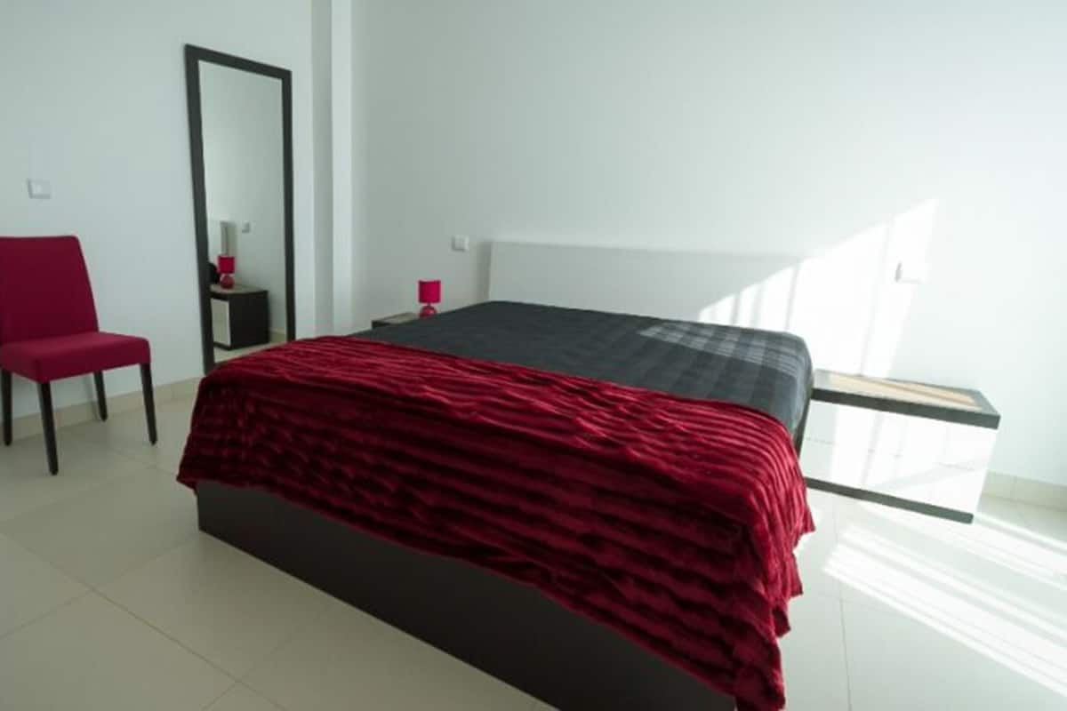 A bedroom with a bed, a chair, a mirror, and a bedside table.
