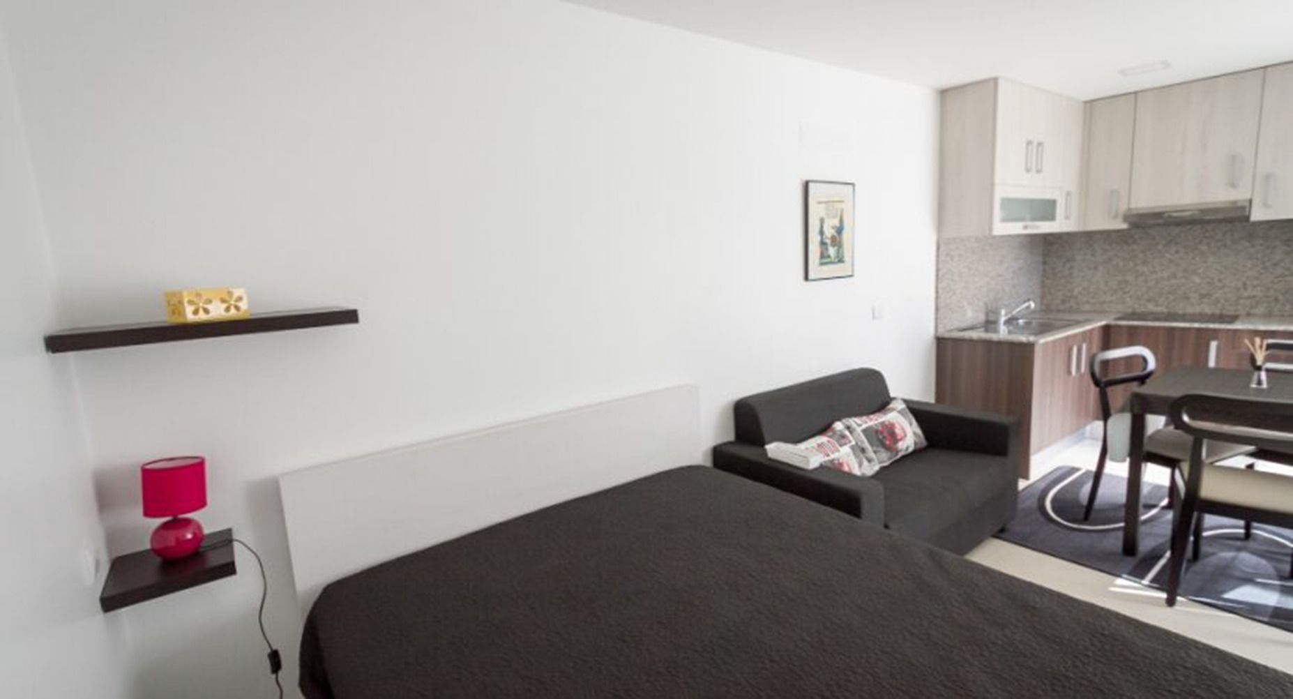 A compact room with a bed, a small sofa, a dining table, and a kitchenette.