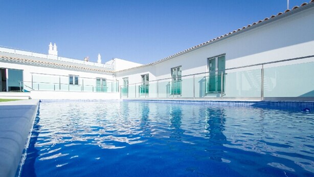Una piscina con acqua cristallina azzurra, circondata da un edificio bianco con ampie finestre e un tetto in tegole.
