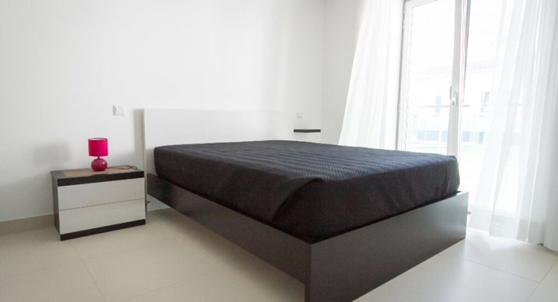 Een slaapkamer met een bed, nachtkastje, lamp en raam met gordijnen.