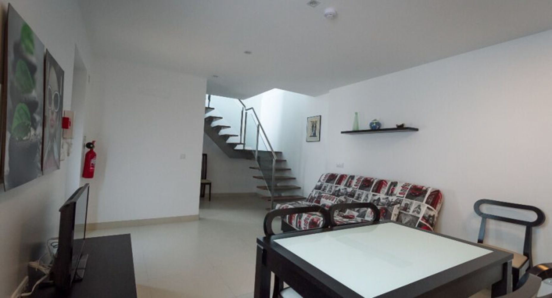 A living room with a sofa, a dining table, a TV, and a staircase leading to another room.