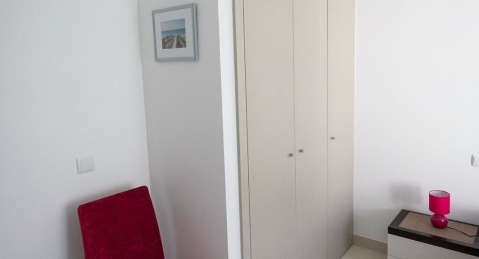 A room with a red chair, a white wardrobe, and a small table with a pink lamp.