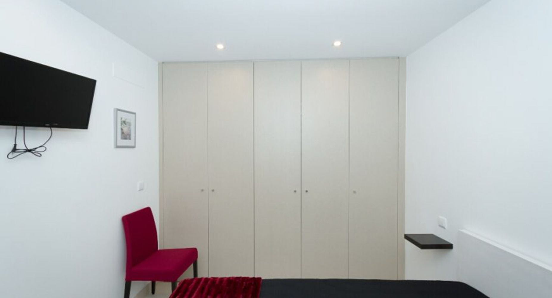 Um quarto com cama, uma cadeira vermelha, um guarda-roupa, uma TV fixada na parede e uma pequena estante.