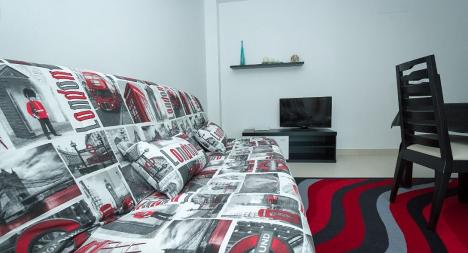 A modern living room with a sofa covered in a newspaper print, a black TV stand, a black chair, and a red and black rug.