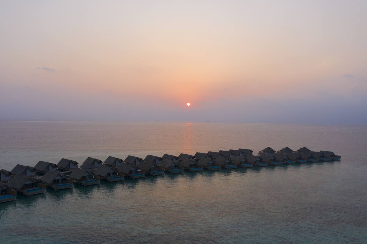 Wasserbungalows reihen sich aneinander, während die Sonne im Hintergrund untergeht.