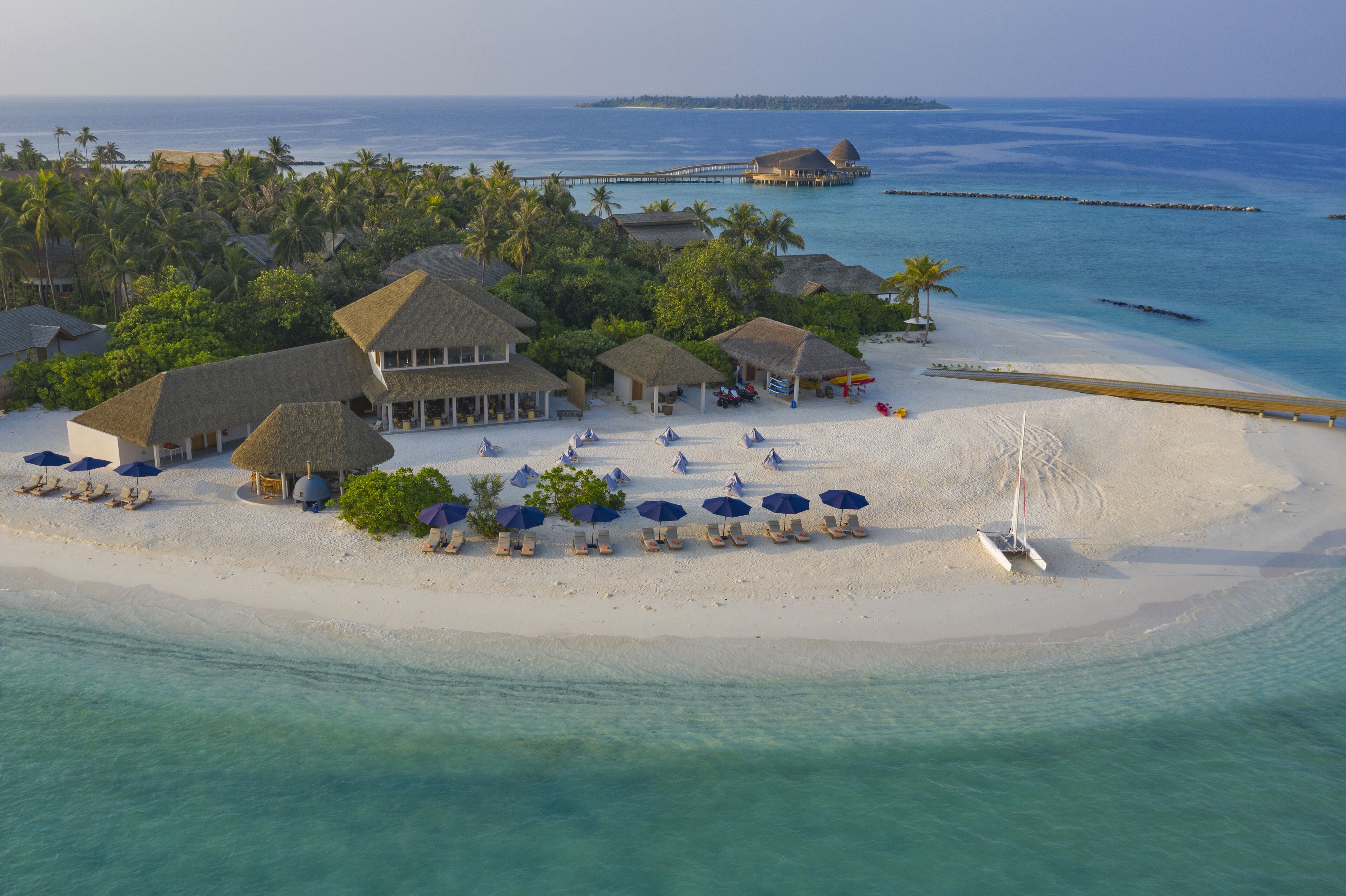 An aerial view of a beach resort with thatched-roof buildings, sun loungers, and umbrellas.